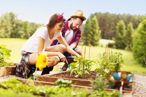 A young couple, a woman with long brown hair and a man wearing a straw hat, are working together in a garden bed in a backyard. The woman is wearing glasses, a white t-shirt, and plaid shorts, while the man is dressed in a plaid shirt and jeans. They appear to be tending to vegetable plants, with several small plants and seedlings visible in the raised garden bed, which is bordered by wooden planks. The garden features rich, dark soil, and the surrounding area includes a neatly maintained lawn with lush green grass, a few decorative garden ornaments, and a stone pathway. In the background, there are tall, leafy trees and a bright, clear sky, indicating a sunny day. This outdoor setting showcases a well-maintained garden space that supports gardening activities, consistent with landscaping services offered by Landscapers Twickenham, in an area near Twickenham or the TW postcode district. The scene reflects a peaceful, productive planting environment ideal for outdoor gardening and yard maintenance.