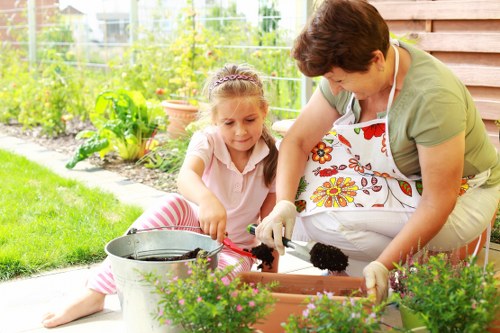 In a backyard garden, a woman and a young girl are engaged in gardening activities. The woman, wearing a floral apron and gardening gloves, is tending to a rectangular flower bed filled with dark, moist soil, while the girl, dressed in a light pink top and striped pants, is assisting by holding a small garden trowel. The garden features lush green grass and a variety of vibrant, blooming plants in the foreground, with some potted plants and a bucket nearby. In the background, there is a wooden fence and a well-maintained vegetable garden with leafy green plants and small shrubs. The scene is set in bright daylight with natural sunlight highlighting the textures of the plants and soil, indicating a pleasant outdoor environment suitable for gardening services in Twickenham or nearby areas. This image visually represents typical outdoor gardening and planting activities that a professional landscaper might provide, including planting, soil preparation, and maintaining a healthy garden space.
