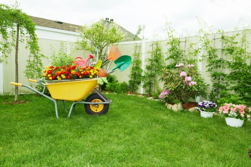 A well-maintained residential garden featuring a lush, evenly cut green lawn that covers the majority of the outdoor space. In the foreground, a yellow garden wheelbarrow is filled with vibrant flowering plants, including red, yellow, and orange blossoms, positioned on the grass. Behind the wheelbarrow, a variety of garden tools such as a green watering can and an orange watering bucket are resting against a small shrub. To the right, several white pots and flower beds contain pink, purple, and white spring-flowering plants, adding colour to the landscape. The garden is enclosed by a white wooden fence, behind which climbing plants and small trees are growing, providing a natural backdrop. The environment appears bright and clear, suggesting a sunny day, suitable for outdoor gardening activities. This scene reflects typical lawn and garden maintenance in Twickenham or nearby areas, offered by professional services like Landscapers Twickenham, emphasizing a tidy, healthy, and inviting outdoor space connected to local gardening standards.
