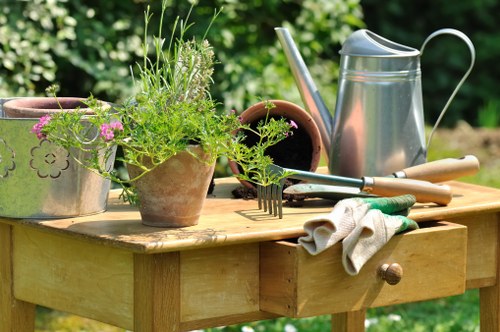 A wooden garden table outdoors, situated in a lush green garden with trees and shrubs in the background, displays various gardening tools and supplies including a metal watering can, a pair of gardening gloves, and multiple terracotta pots with some containing live plants with green foliage and pink flowers. The table's surface is polished, with a small drawer visible on the front side, and the scene suggests preparation or ongoing gardening activities typical of a residential outdoor space in Twickenham, with natural daylight highlighting the vibrant greens and earthy tones and emphasizing the connection between outdoor maintenance and professional gardening services.