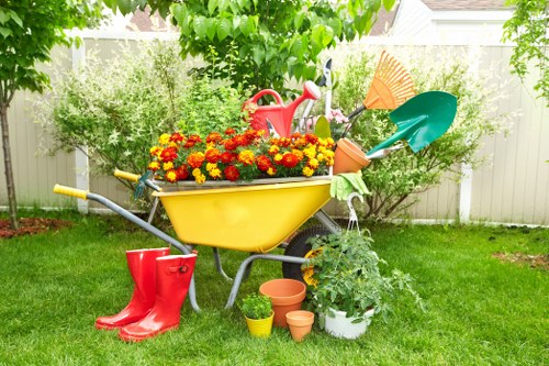 A vibrant garden scene featuring a central yellow wheelbarrow filled with red, orange, and yellow flowering plants, positioned on a well-maintained green lawn. Surrounding the wheelbarrow are garden tools including a red watering can, a green spade, and a terracotta pot. To the left, a pair of red rubber boots rests on the grass. Nearby, several small terracotta and plastic plant pots are placed on the ground. In the background, a white fence borders the garden, with lush green shrubs and tall trees providing shade and natural enclosure. Bright sunlight illuminates the scene, indicating a clear day, and a few scattered leaves from the trees add to the natural outdoor atmosphere. The overall setting reflects a well-kept, vibrant outdoor space suitable for gardening activities, with elements that support gardening and outdoor maintenance services like those provided by Landscapers Twickenham.