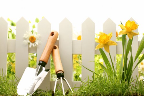 A close-up view of a small garden border featuring vibrant yellow daffodils and white daisies growing alongside a white wooden picket fence, which provides a neat and decorative boundary for the garden. The foreground shows freshly planted green grass and rich, dark soil, indicating recent planting or soil preparation. Resting on the soil are gardening tools including a hand trowel with a light wooden handle and a small gardening fork, suggesting ongoing garden maintenance or flower bed creation by Landscapers Twickenham. The background reveals soft, natural daylight, likely during springtime, highlighting the lush greenery and bright flowers against the fence, with hints of blurred foliage. This outdoor space reflects a well-maintained garden area suitable for gardening services such as flower bed creation, planting, and outdoor landscape management within the Twickenham area, demonstrating a neat and vibrant garden environment ready for further landscaping enhancements.