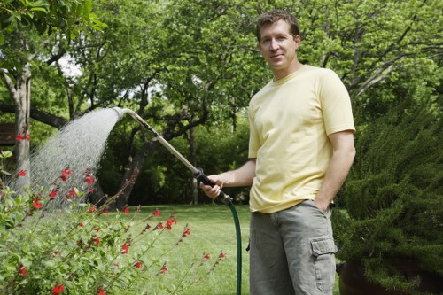 A man wearing a light yellow t-shirt and grey cargo shorts stands in a well-maintained garden, watering flowering plants with a garden hose. The garden features a lush green lawn, flowering shrubs with red blossoms, and neatly trimmed bushes. Tall trees with leafy canopies provide shade in the background, along with a dense hedge on the right side, all set under a partly cloudy sky. The scene suggests outdoor maintenance activity typical in residential gardens within Twickenham, aligning with landscaping and gardening services offered by Landscapers Twickenham. The environment appears vibrant and healthy, with natural tones of green, red, and earthy soil visible near plant beds, demonstrating a cared-for outdoor space suitable for garden improvements or routine lawn care.