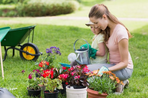 In a well-maintained garden, a woman and a young girl are engaged in planting flowers or working with garden pots on a lush, green lawn. The foreground features an array of colourful flowering plants in black, white, and terracotta pots, with vibrant red, pink, purple, and orange blossoms. Behind them, the lawn appears dense and healthy, bordered by a neatly trimmed hedge, providing a sense of enclosure and privacy typical of suburban gardens in Twickenham. To the side, a garden wheelbarrow with yellow wheels is positioned on the grass, indicating ongoing outdoor maintenance or planting activity. The background showcases a softly blurred garden environment with a sense of openness and natural light, suggesting a clear day with mild weather conditions. The scene subtly reflects typical gardening services offered by Landscapers Twickenham, emphasizing planting, lawn care, and outdoor maintenance aspects of a well-kept garden environment, situated within the TWickenham area. The overall setting is relaxed and meticulously cared for, highlighting professional gardening and landscaping work that enhances outdoor spaces in the local region.
