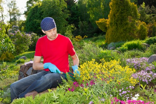 A young man wearing a blue patterned gardening hat, red t-shirt, and light blue gloves is sitting amidst a well-maintained garden in Twickenham, with lush greenery and a variety of flowering plants. The garden features a diverse mix of colourful flowers, including yellow, pink, and purple blooms, arranged in naturalistic flower beds and borders. Dense green shrubs and small trees form a backdrop behind the vibrant flower display, creating a peaceful outdoor space typical of landscaped gardens in the Twickenham area. The scene is captured during daylight with natural sunlight illuminating the garden, highlighting the textures of foliage, soil, and paving. The overall setting suggests a carefully designed garden environment, ideal for outdoor leisure or gardening maintenance, aligning with professional gardening services offered by Landscapers Twickenham to enhance outdoor spaces in local areas.