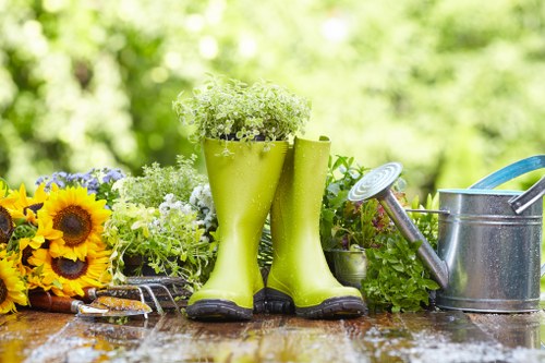 A garden scene featuring a wooden outdoor table with various garden tools and decorative elements, including a pair of bright green rubber gardening boots filled with small white flowers, placed next to a silver metal watering can with a blue handle. To the left, there is a vibrant arrangement of sunflowers with yellow petals and dark centers, alongside potted plants and greenery. The background displays a lush, out-of-focus green garden with sunlight filtering through trees, creating a bright and inviting outdoor atmosphere suitable for gardening activities and landscape care services in Twickenham, UK.