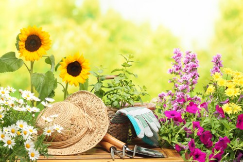 A vibrant garden scene featuring a variety of flowering plants and gardening tools set on a wooden surface. The foreground displays bright yellow sunflowers with large, dark centers, alongside white daisies with yellow centers and clusters of purple and pink flowers, all surrounded by green foliage. A wide-brimmed straw hat with a decorative band rests atop the wooden surface, next to gardening gloves, a small trowel, and pruning shears, suggesting recent or ongoing garden maintenance. The background reveals a lush, green outdoor space with softly blurred trees and foliage, indicative of a sunny day with natural light illuminating the scene. The overall arrangement reflects an inviting, well-maintained outdoor garden typical of residential landscaping in Twickenham, with a focus on floral diversity and garden care tools, subtly supporting gardening services offered by Landscapers Twickenham to enhance different outdoor spaces and promote garden health and aesthetics.