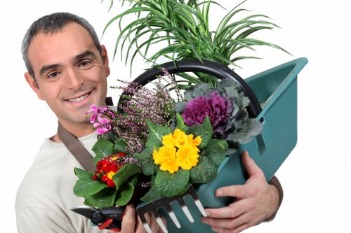 The image depicts a smiling man holding a small teal plastic garden watering can filled with a variety of potted plants and flowers, including yellow, purple, pink, and red flowering plants with green foliage. The background is plain, emphasizing the garden elements and the man's friendly expression. This scene relates to gardening and outdoor plant care, aligning with landscaping services offered by Landscapers Twickenham. The plants appear healthy and vibrant, suitable for home garden enhancement in Twickenham, with the watering can symbolizing outdoor maintenance activities. The overall composition suggests professional gardening support, focusing on plant care, flower arrangement, and shrub or garden bed maintenance typical in residential gardens across the Twickenham postcode area, N22. The lighting suggests a bright, outdoor environment, with natural tones highlighting the vivid colors of the flowers and the lush green foliage, making it an inviting image for garden care and landscaping services.