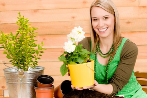 The image shows a smiling woman sitting outdoors in a garden space with a wooden fence in the background. She is holding a yellow flower pot containing a healthy, blooming white flower with green leaves. In front of her on the table, there are additional plant containers, including a large silver metal pot with a small, leafy green shrub and several empty black and red pots ready for planting. The garden area features a well-maintained lawn with dense, dark green grass, and the overall setting appears to be a neatly landscaped backyard or front garden intended for gardening activities. The weather seems bright and clear, with natural daylight illuminating the scene, emphasizing the lush greenery and vibrant flowers. This setup reflects professional gardening and landscaping services, such as flower bed creation and outdoor plant care, offered by Landscapers Twickenham, aligning with the services detailed on the Twickenham Flower Bed Services webpage.