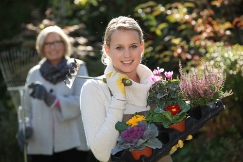 A young woman with blonde hair, wearing a white sweater and gardening gloves, is standing in a well-maintained garden during daylight, holding a tray of vibrant flowering plants with purple, pink, yellow, and red blooms. Behind her, an older woman with glasses, a light-colored jacket, and a scarf is smiling and holding garden pruning tools, suggesting outdoor garden maintenance activities. The background features lush green foliage and trees with sunlight filtering through, highlighting the natural tones of the garden. The scene emphasizes gardening work, with a diverse mix of plants, soil beds, and garden tools visible, reflecting a typical outdoor space that could benefit from professional gardening services offered by Landscapers Twickenham in the local area near Twickenham, TW1 postcode, aligning with the service focus on landscaping and garden care.